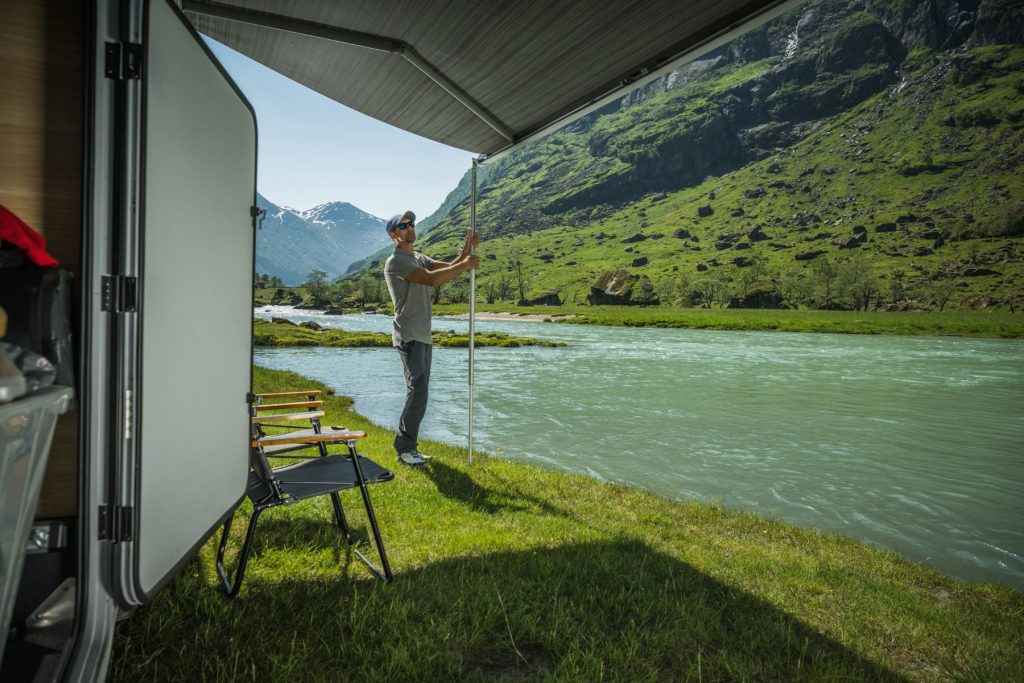 Man sets up his travel trailer's awning at his off-grid campsite next to a stream
