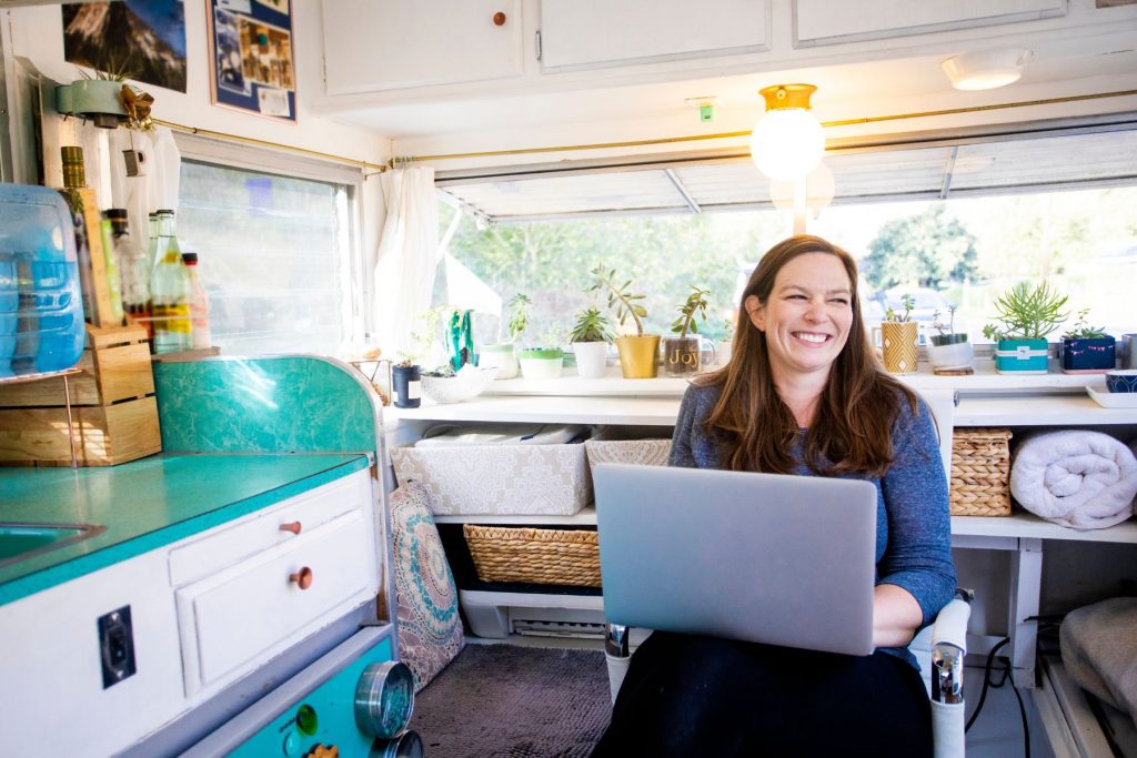 Woman sits in her travel trailer with her laptop open, smiling.