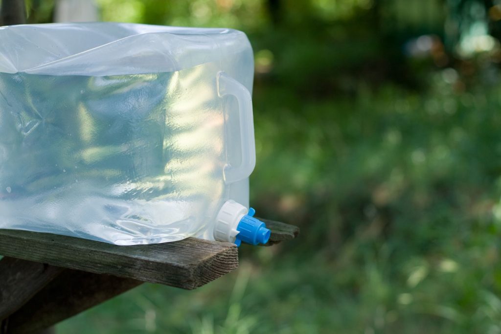 Close up of a collapsible clear water container resting on the side of a picnic table at a campsite.